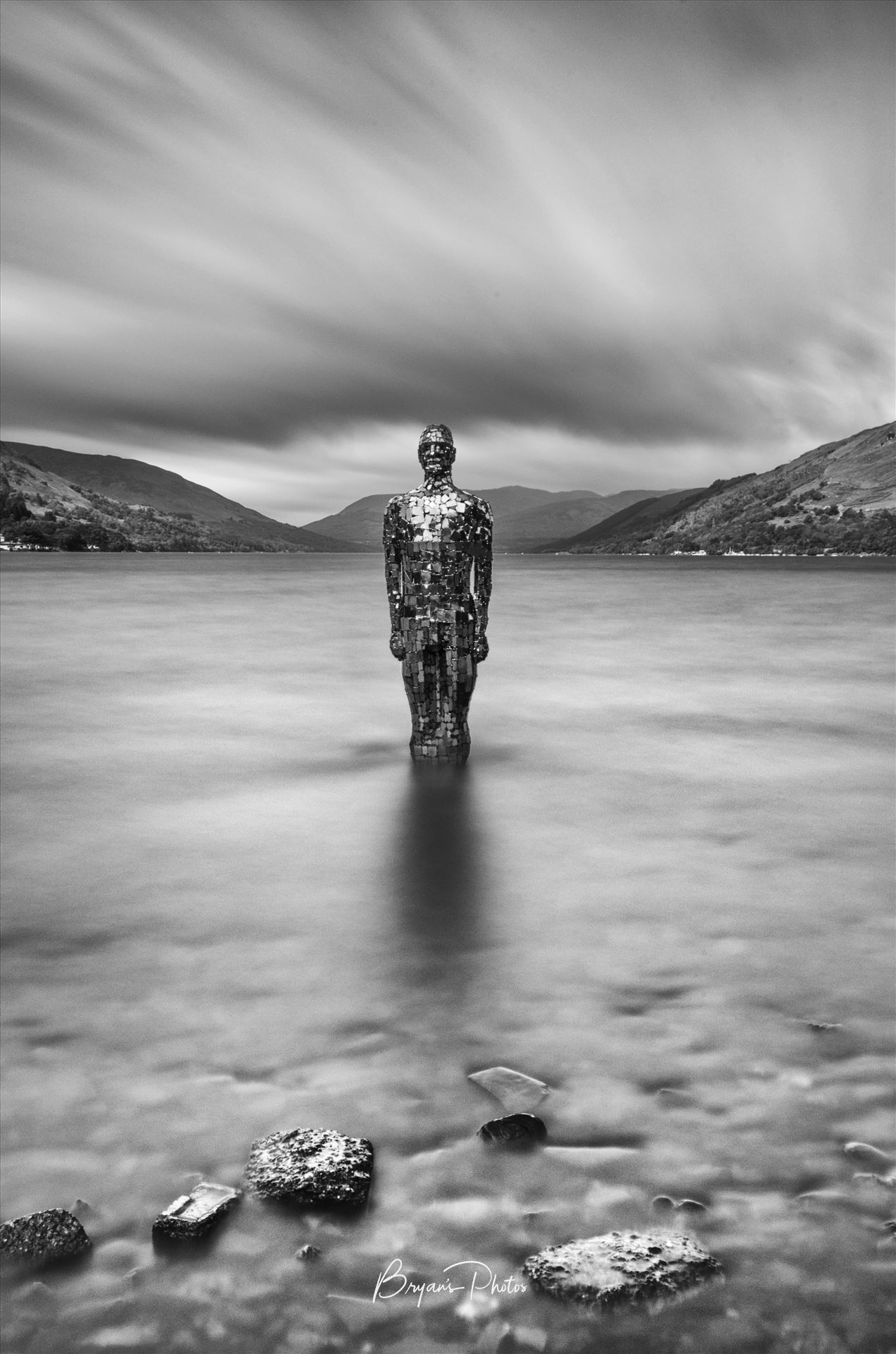 Mirror Man - A black and white long exposure photograph of the still mirror man sculpture taken from St Fillans at Loch Earn. by Bryans Photos