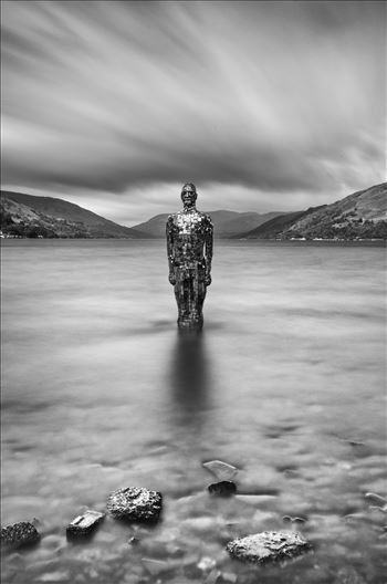 A black and white long exposure photograph of the still mirror man sculpture taken from St Fillans at Loch Earn.